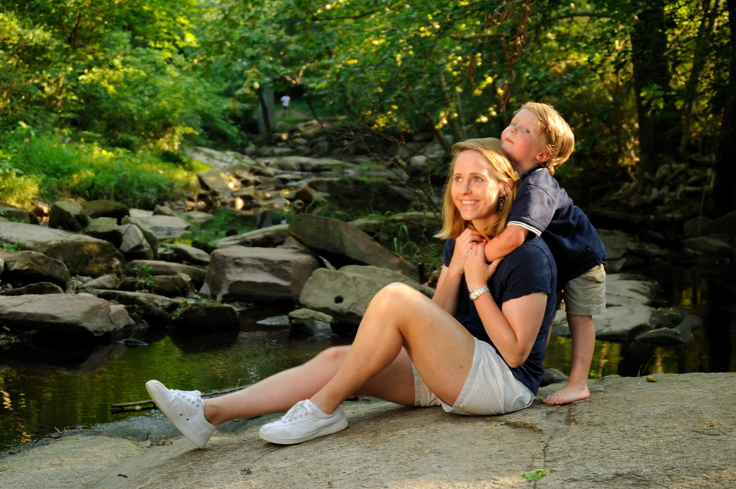 A woman and her son sitting on rocks near a stream.