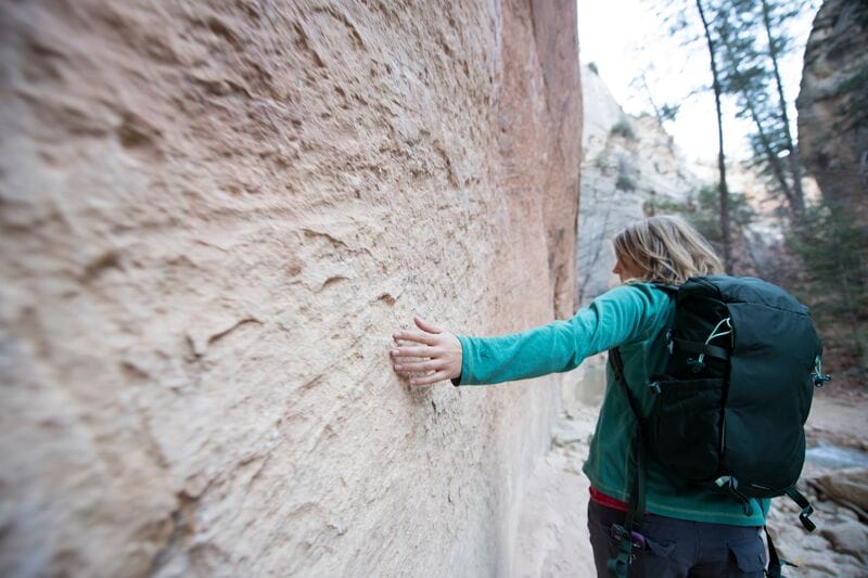 A woman with a backpack reaching out to a rock wall.