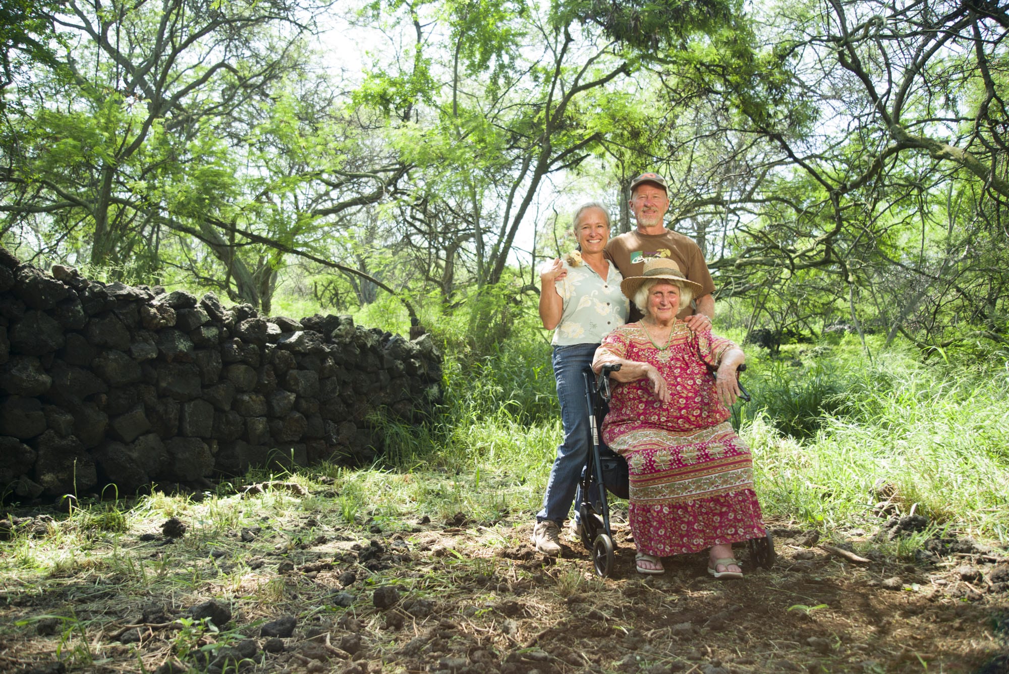 Three people standing next to a stone wall in a wooded area.