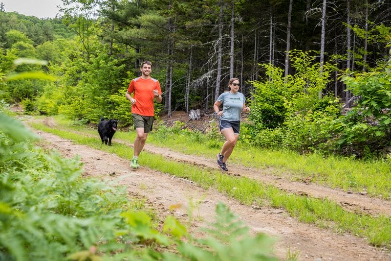 Two people jogging on a dirt road in the woods.