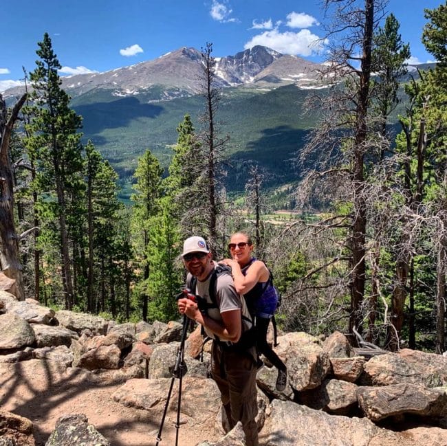 Trevor Hahn and Melanie Knecht smile for a photo on trail in front of a mountain vista. Trevor carries Melanie on his back.