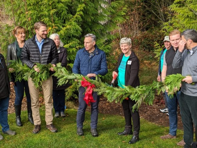 About 10 people in a green lawn hold a long garland and prepare to cut it with scissors