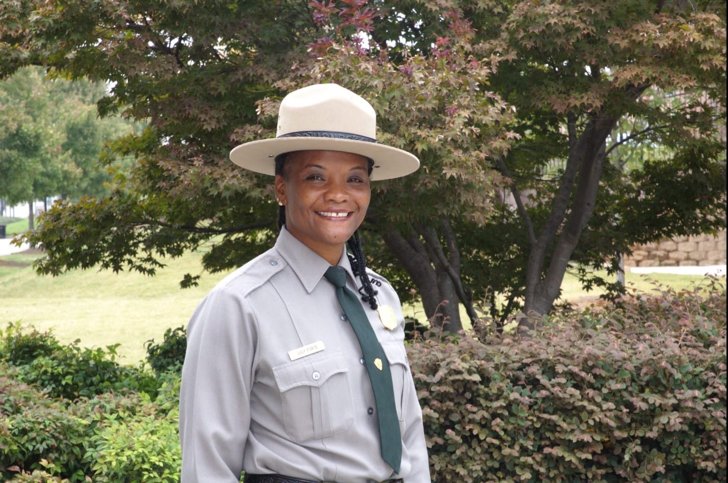 A woman wearing a hat and uniform standing in front of bushes.