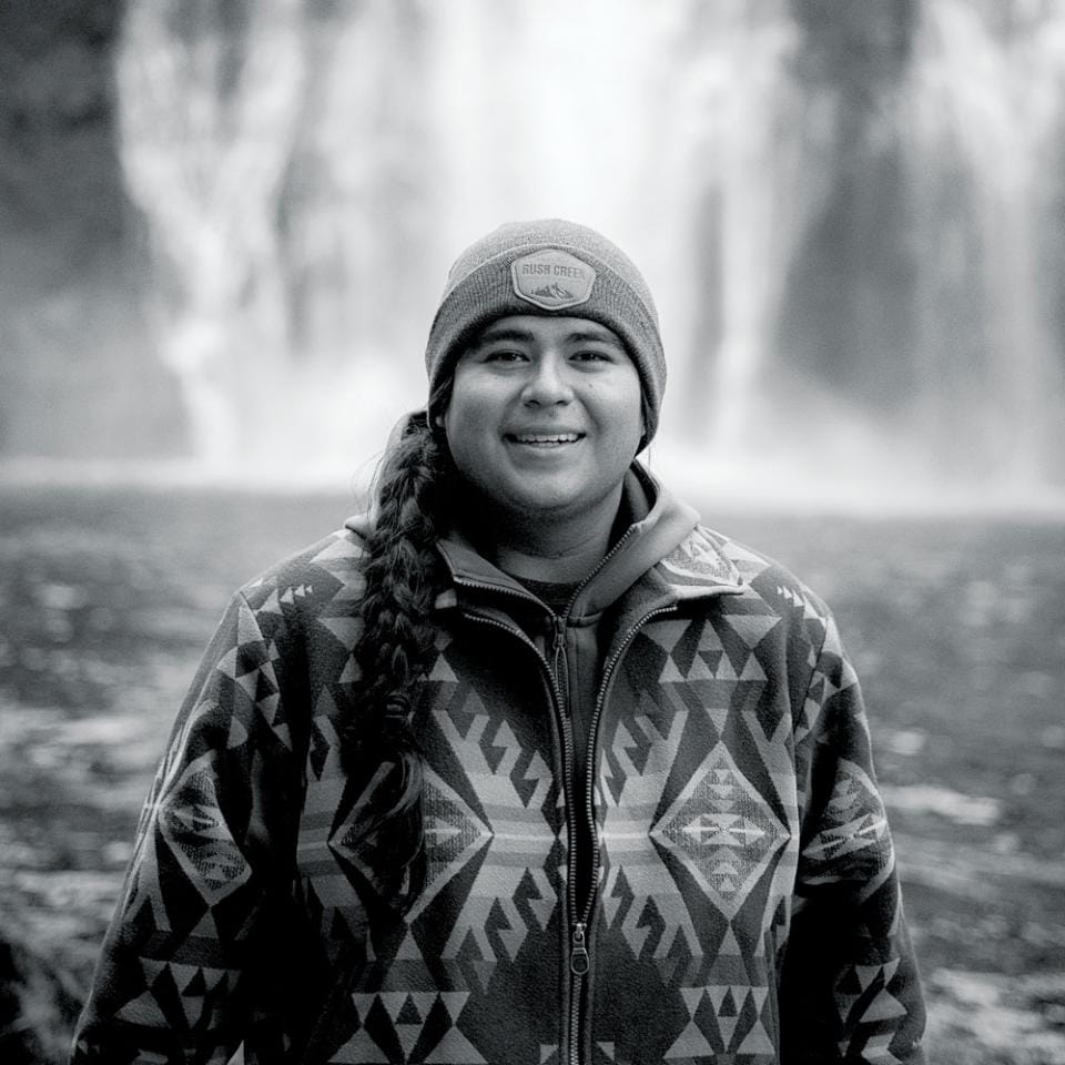 A black and white photo of a woman standing in front of a waterfall.