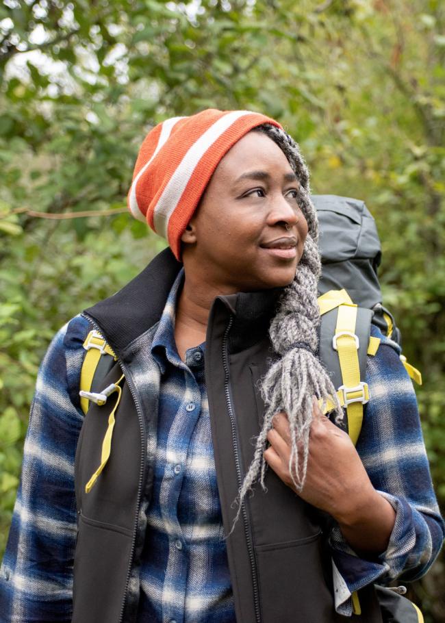 A black woman wearing a plaid shirt and a beanie in the woods.