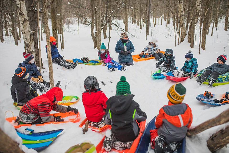 A group of children sitting on snow sleds in the woods.