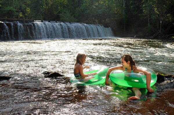 Two girls are floating in an inflatable ring in front of a waterfall.