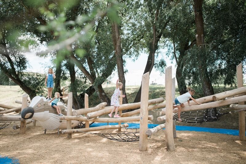 Children playing on a wooden play structure in a wooded area.