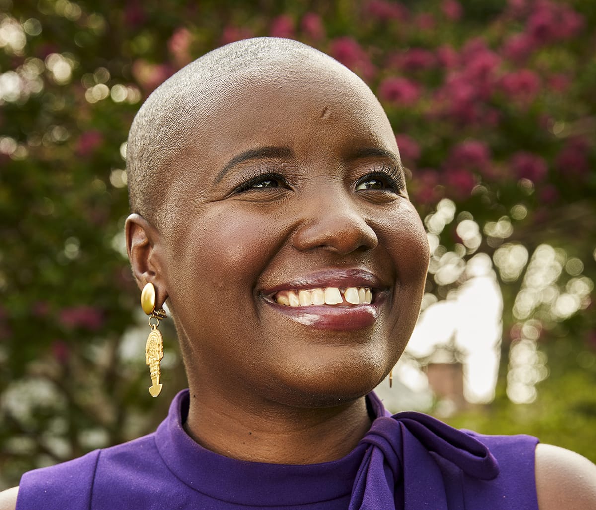 A woman with a shaved head smiling in a purple dress.