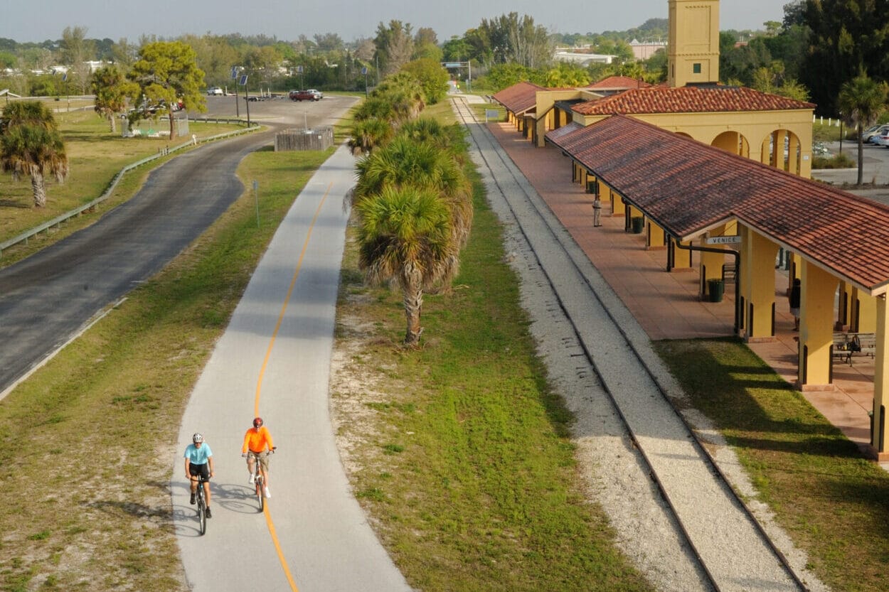 A high view of two bicyclists riding past an yellow train station in Florida, with rail lines parallel to the paved bike trail.