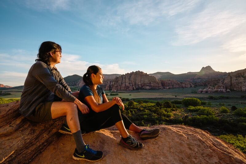 A man and woman sit on top of a rock overlooking a valley.