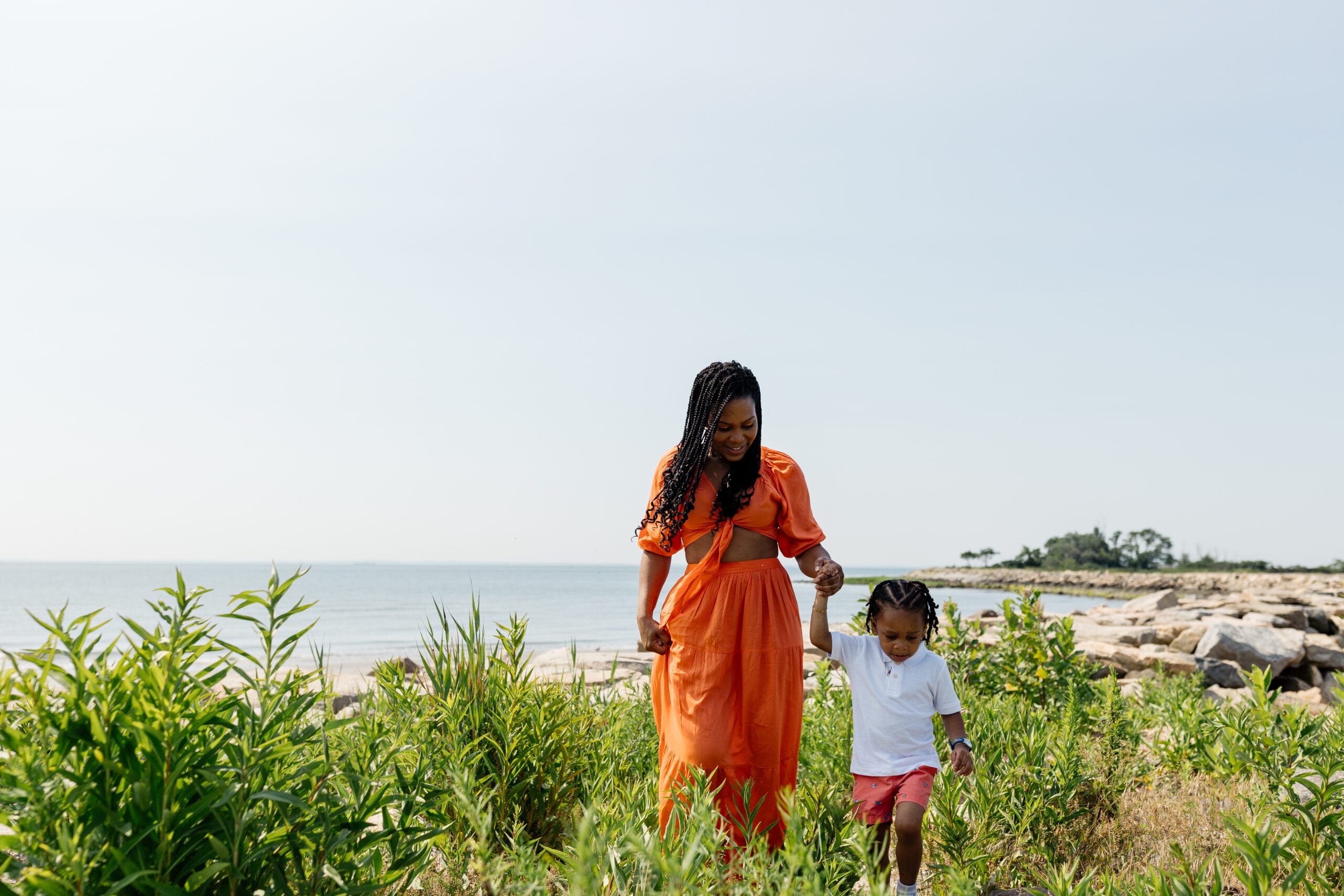 A woman in an orange dress and a child walking through tall grass near the ocean.