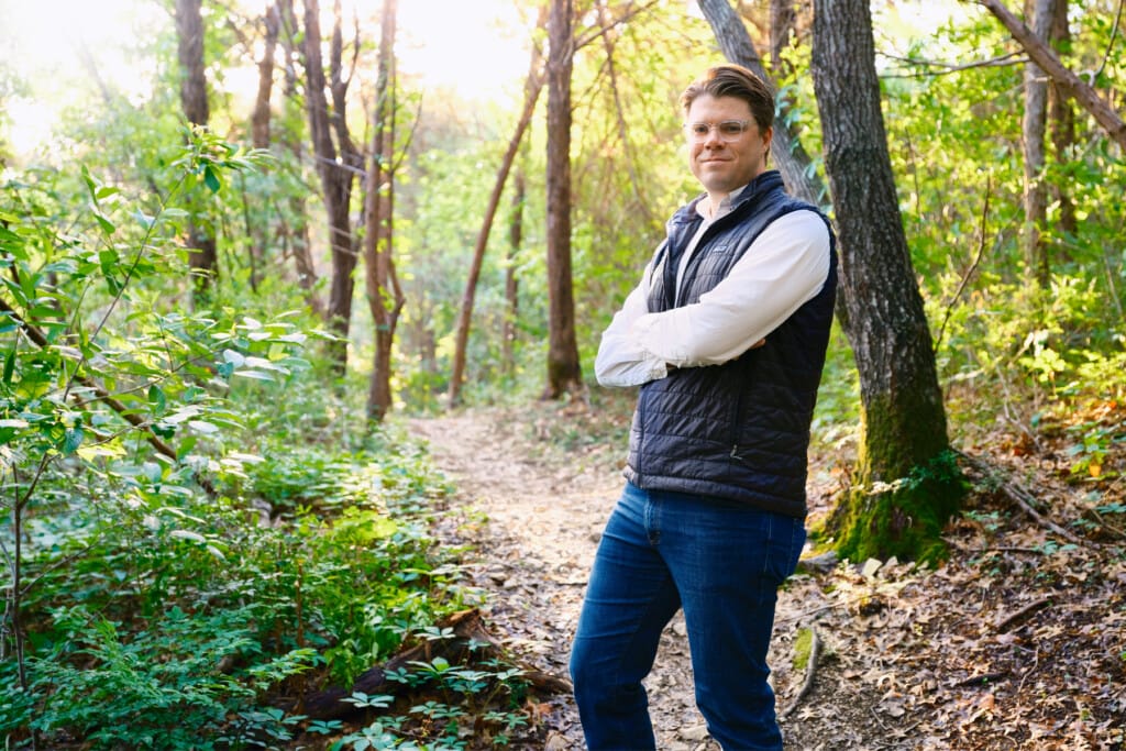 A man in a vest standing on a trail in the woods.