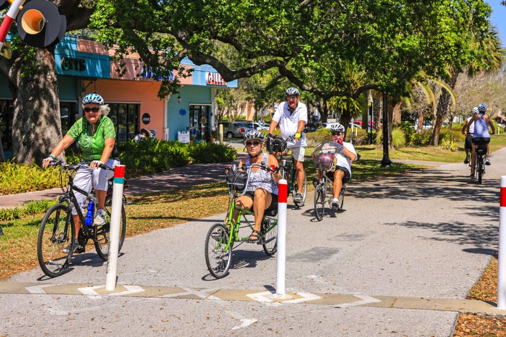 Two women and two men ride upright and recumbent bicycles on a paved trail through a tree-lined commercial area in Florida.