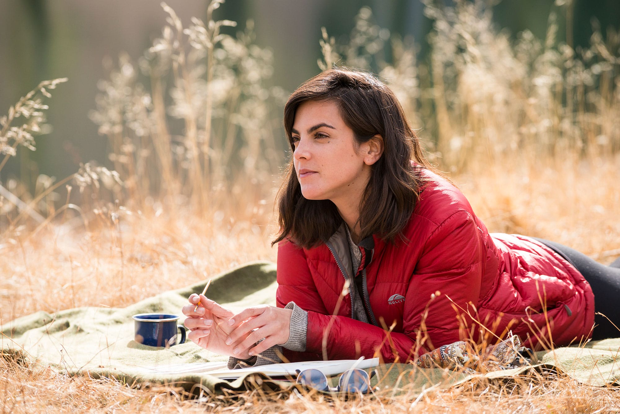 A woman in a red jacket laying on a blanket in a field.