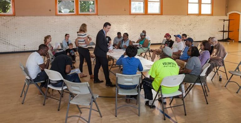 A group of people sit in a circle of chairs in a gymnasium, engaged in discussion with papers on a table in the center.
