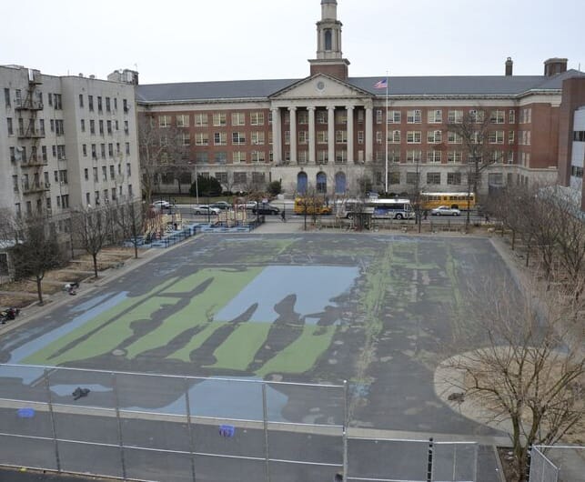 An empty urban schoolyard with faded paint, surrounded by buildings, in front of a large brick school with columns and a flagpole.