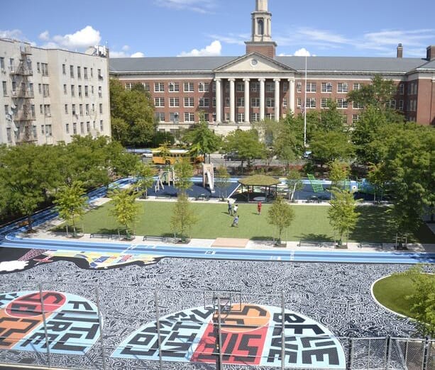 A city school building stands behind a playground with green turf, trees, play structures, and a colorful, patterned mural on the ground in the foreground.