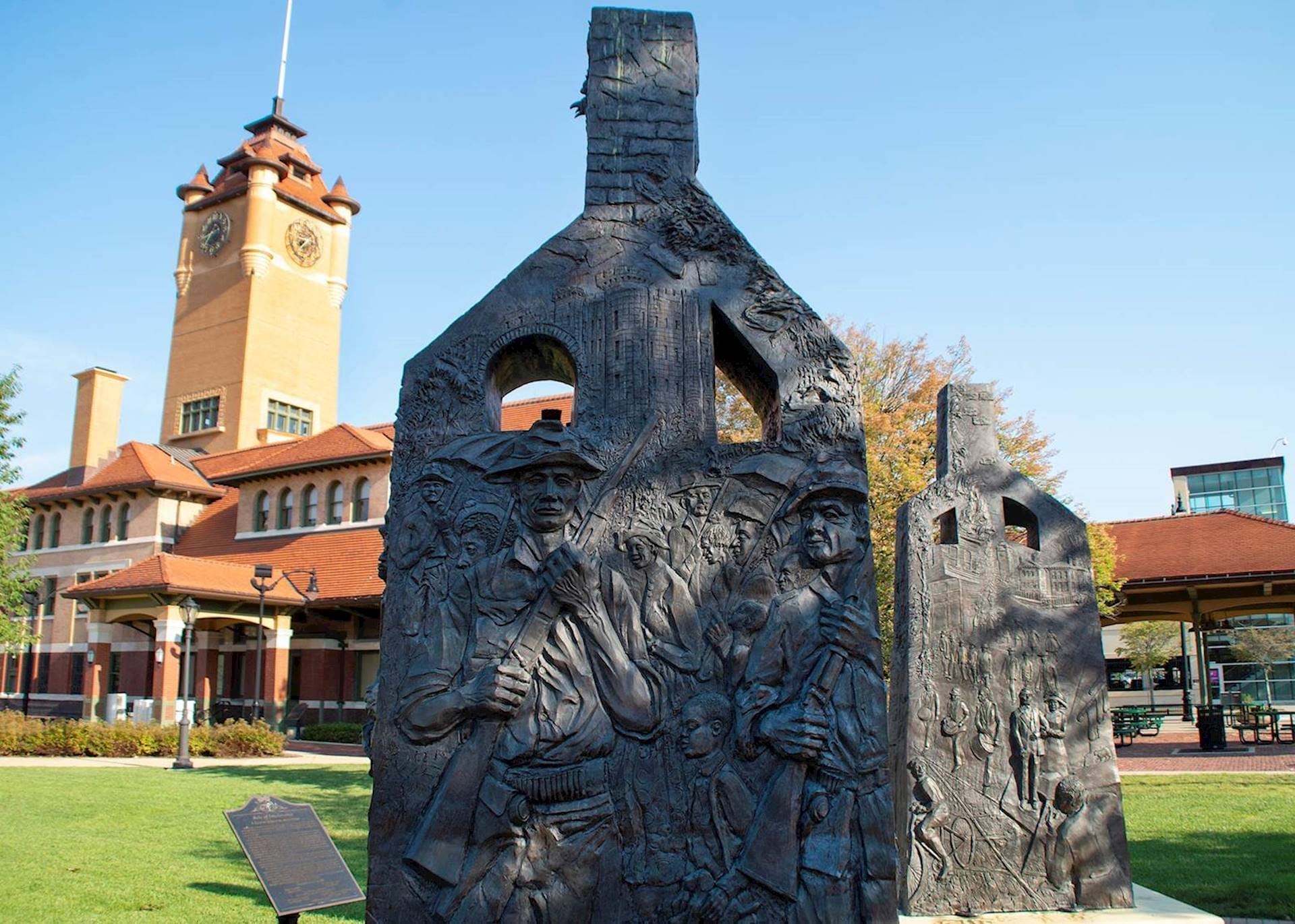 Two chimney-shaped statues engraved with people with a blue sky above and train station in the background