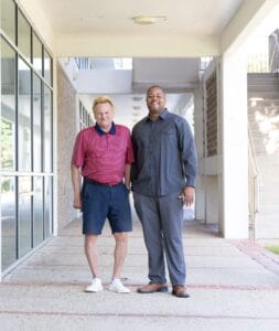 A white man and a Black man stand smiling in an outdoor corridor.