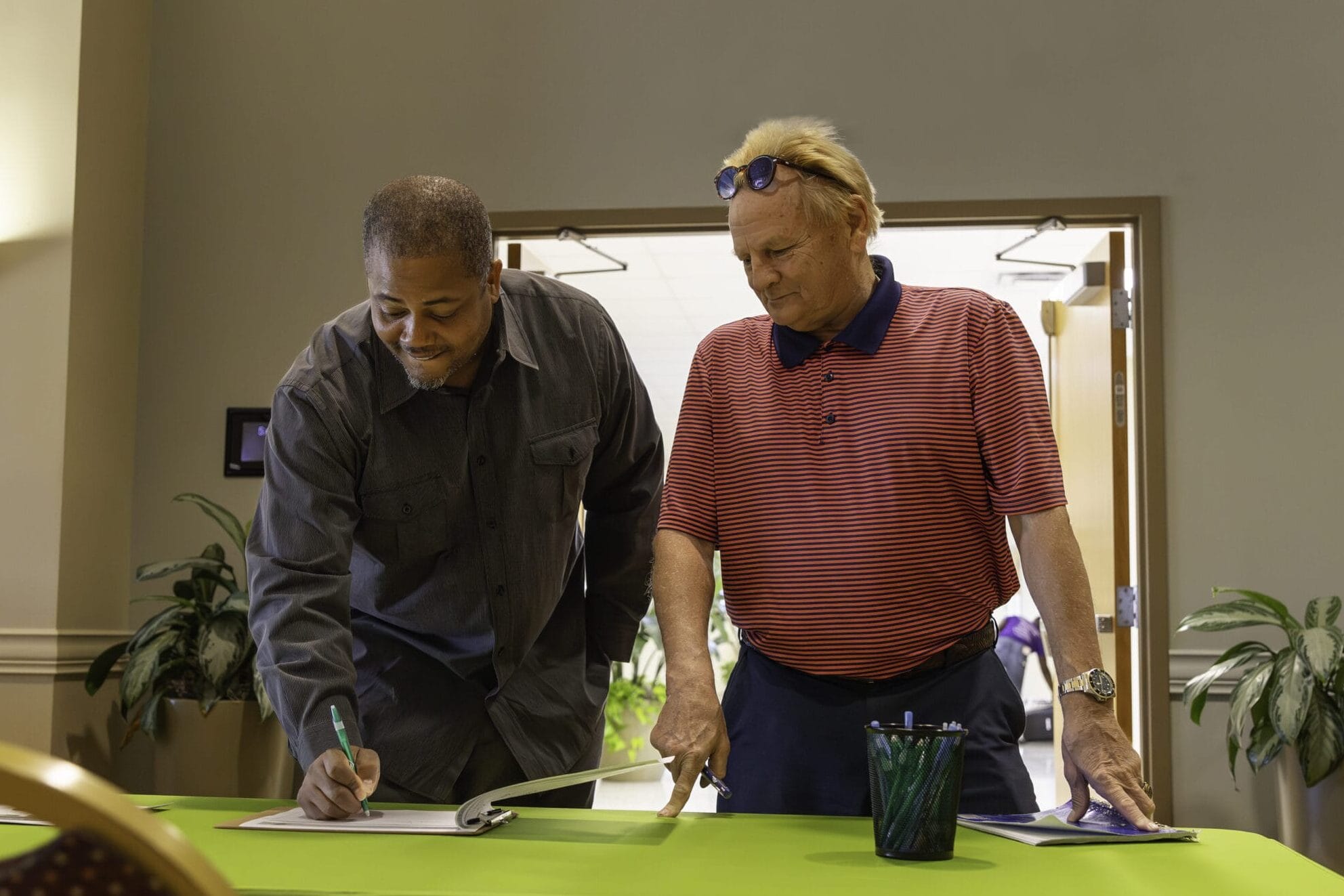 A Black man and a white man stand at a table and look at papers in a binder as if they're planning or working together.