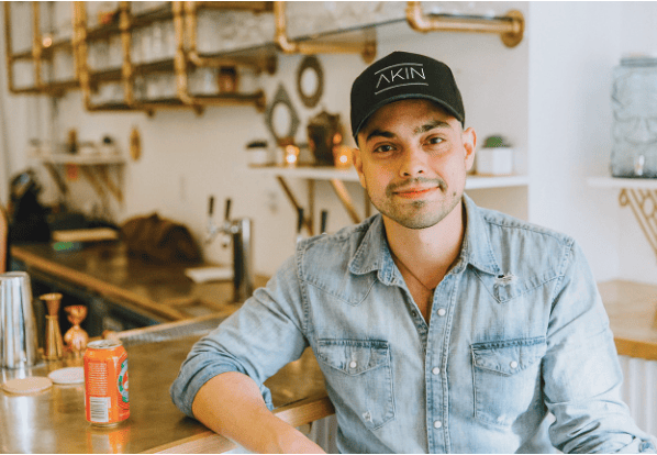 A man in a denim shirt and black cap sits at a counter in a modern café, with a soda can and bar tools visible on the counter beside him.
