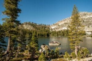 A clear mountain lake surrounded by pine trees and rocky hills under a bright blue sky.