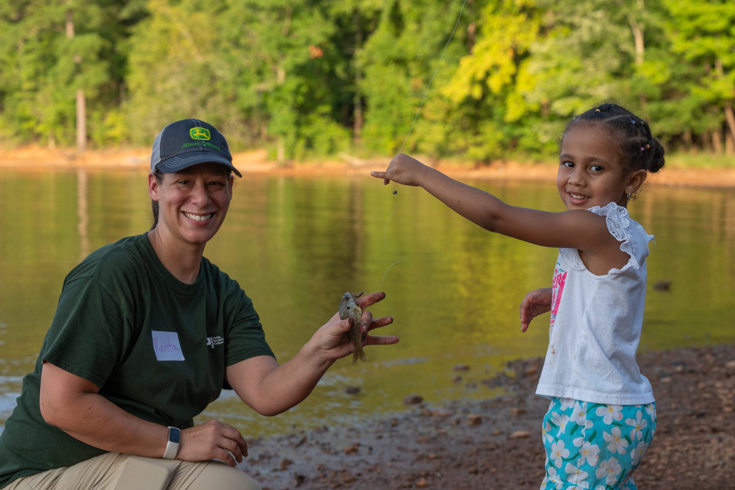 An adult kneels by a lakeshore holding a small fish, while a smiling girl stands beside her pointing at the fish. Trees and water are visible in the background.