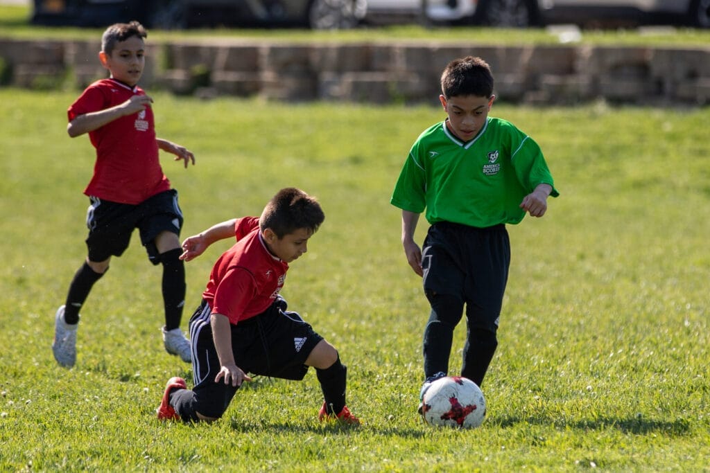 Three young boys play soccer on a grassy field; two in red jerseys defend while one in a green jersey controls the ball.