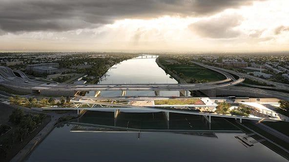 A modern pedestrian bridge with green spaces spans over a river, with highways and urban landscape in the background under a cloudy sky.