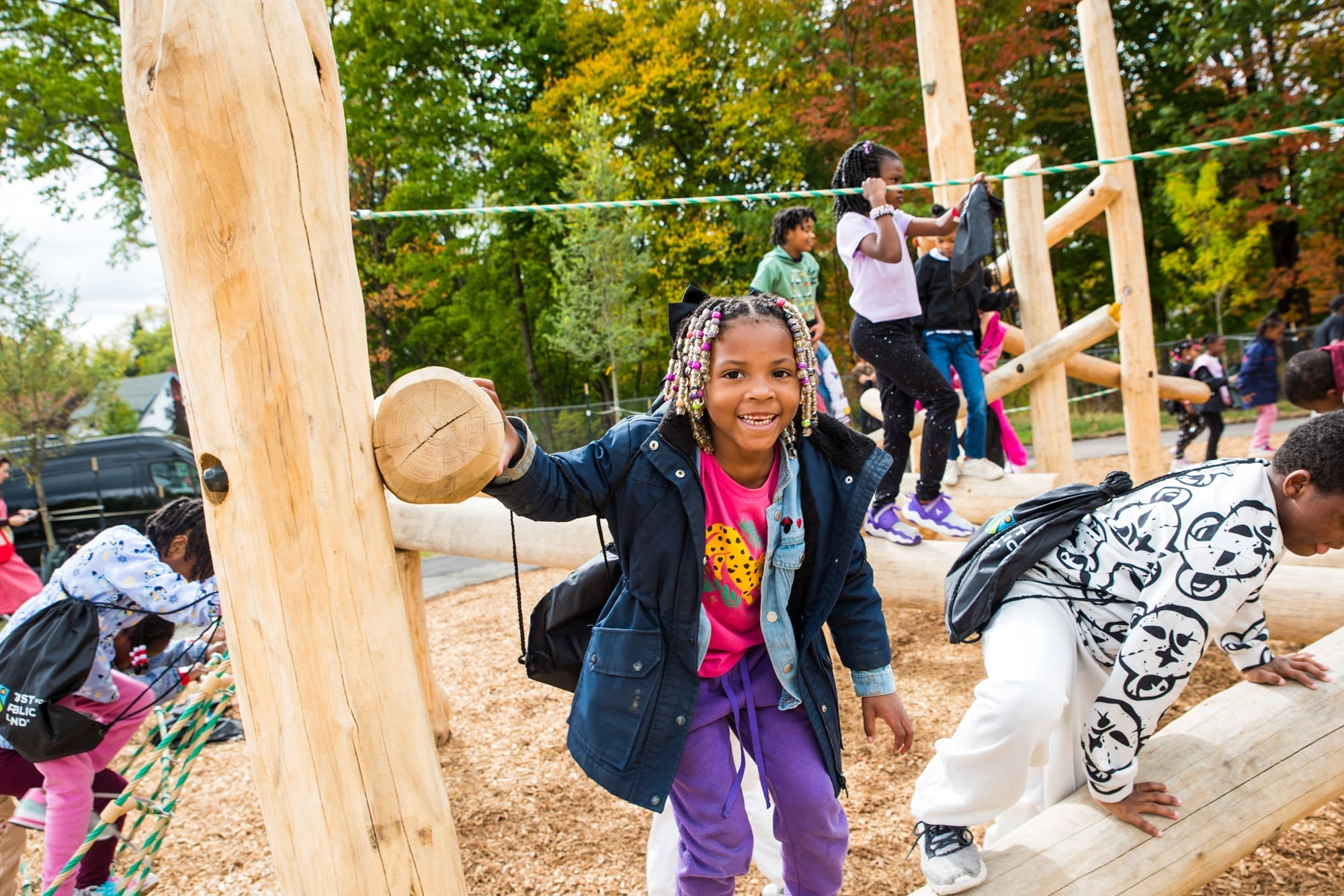 Children climb and play on a wooden playground structure outdoors, surrounded by trees on a sunny day.