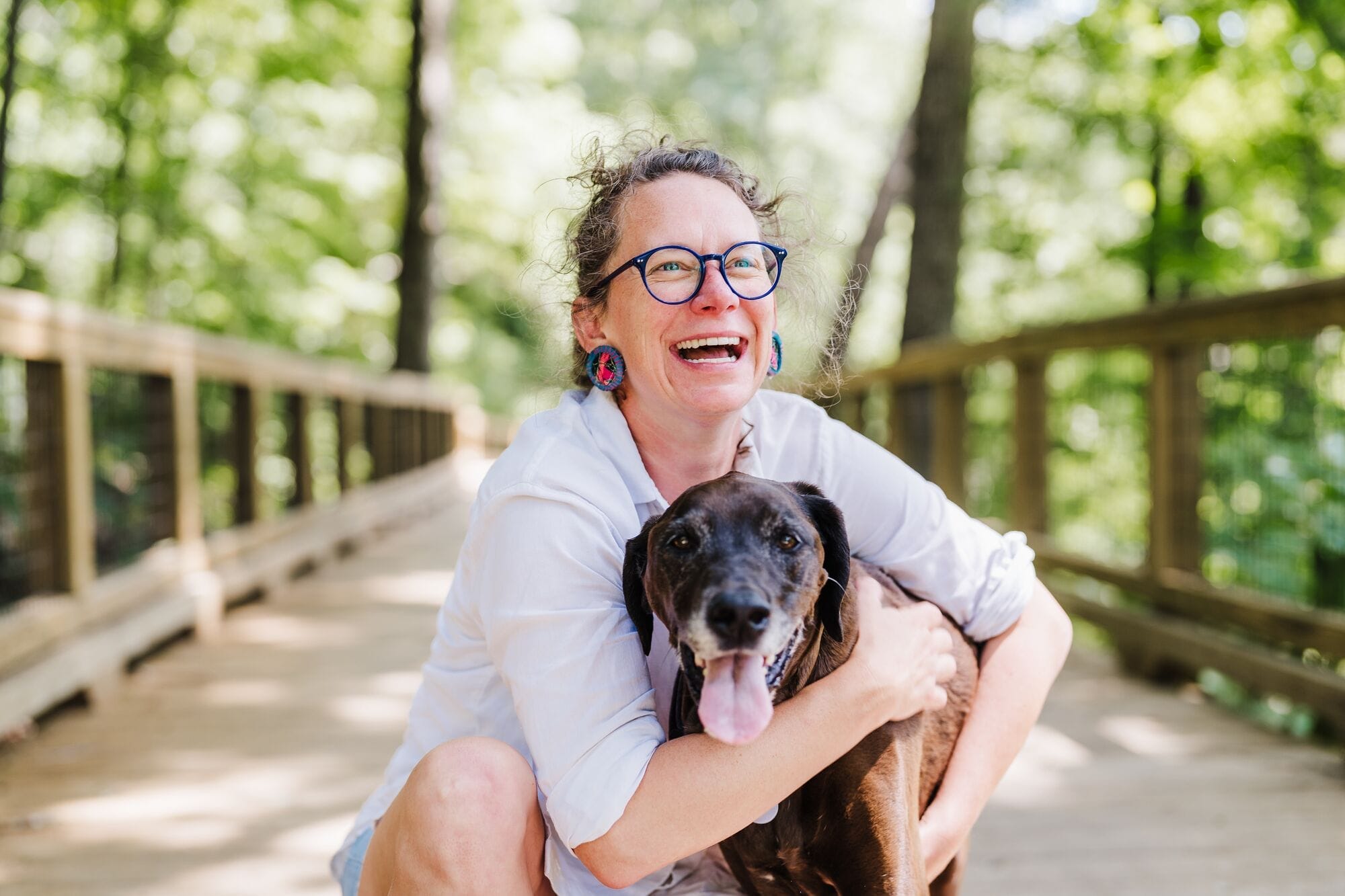 A person wearing glasses and a white shirt kneels on a wooden bridge outdoors, smiling and hugging a black dog with its tongue out.