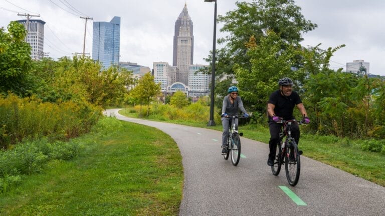 Two people ride bicycles on a paved trail through a green park with a city skyline in the background under a cloudy sky.